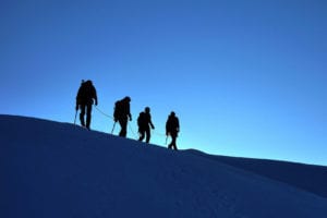 Four mountaineers walk across a snowy ridge.