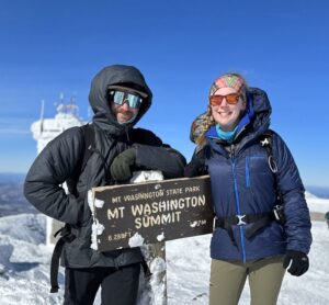 Male and female smile at the summit of Mt. Washington on a sunny day in the winter.