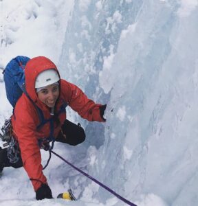 A smiling female in a red jacket ice climbing.