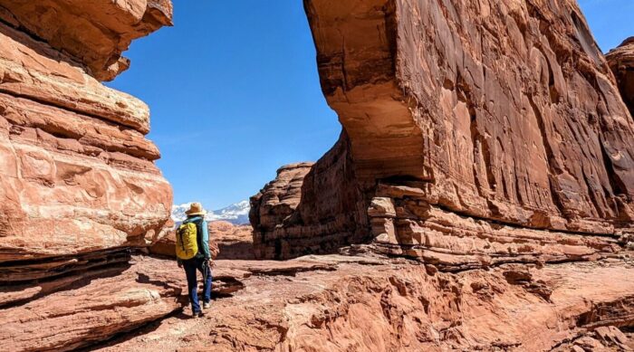 A hiker with a yellow backpack walks under a sandstone arch in Moab, Utah.