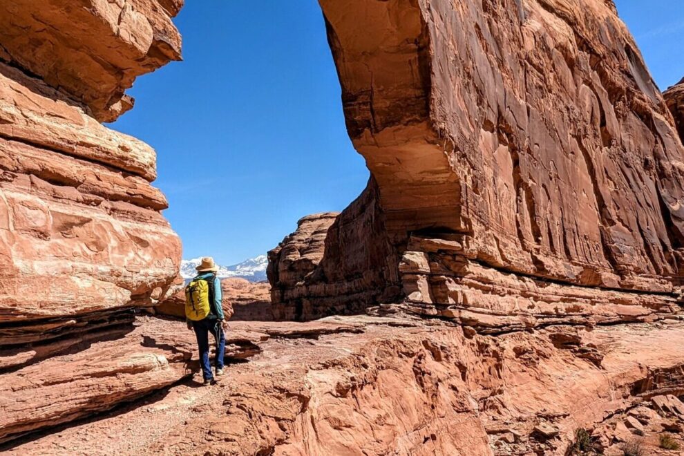 A hiker with a yellow backpack walks under a sandstone arch in Moab, Utah.