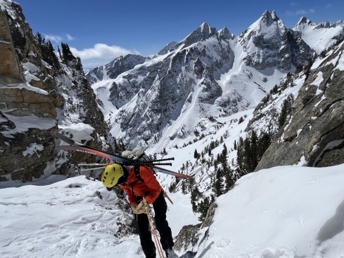 A mountain guide in a yellow helmet rappels into a ski couloir on a sunny day in Grand Teton National Park