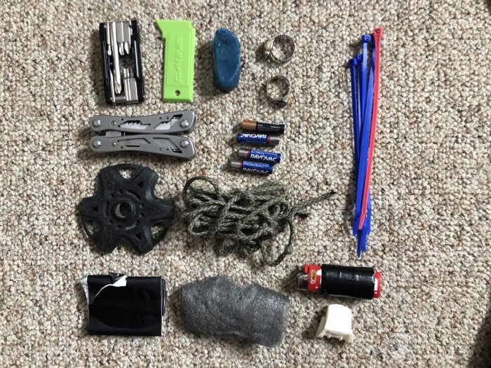 A ski mountaineering repair kit including zip ties, super glue, steel wool, pole basket, hose clamps, binding tool, skin wax, ski straps, paracord, tape, spare batteries, pliers/multi tool, extra skin clips, lighter, gear repair tape, and a scraper